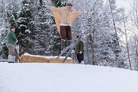 Mann springt mit historischen Ski und Kleidung von einer Mini-Schanze herunter